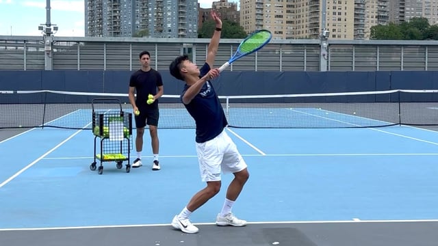 Player serving with a coach and ball cart behind the baseline